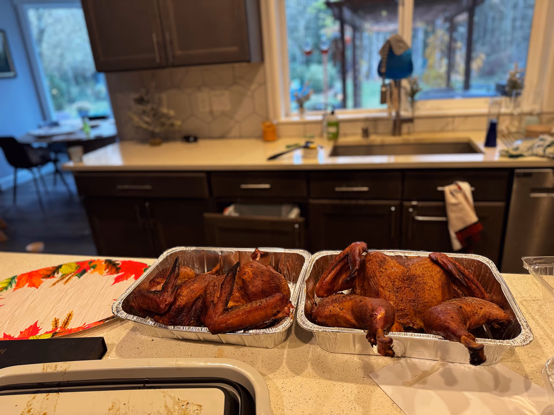 Two aluminum trays of roasted turkey on a kitchen counter with sink and window.