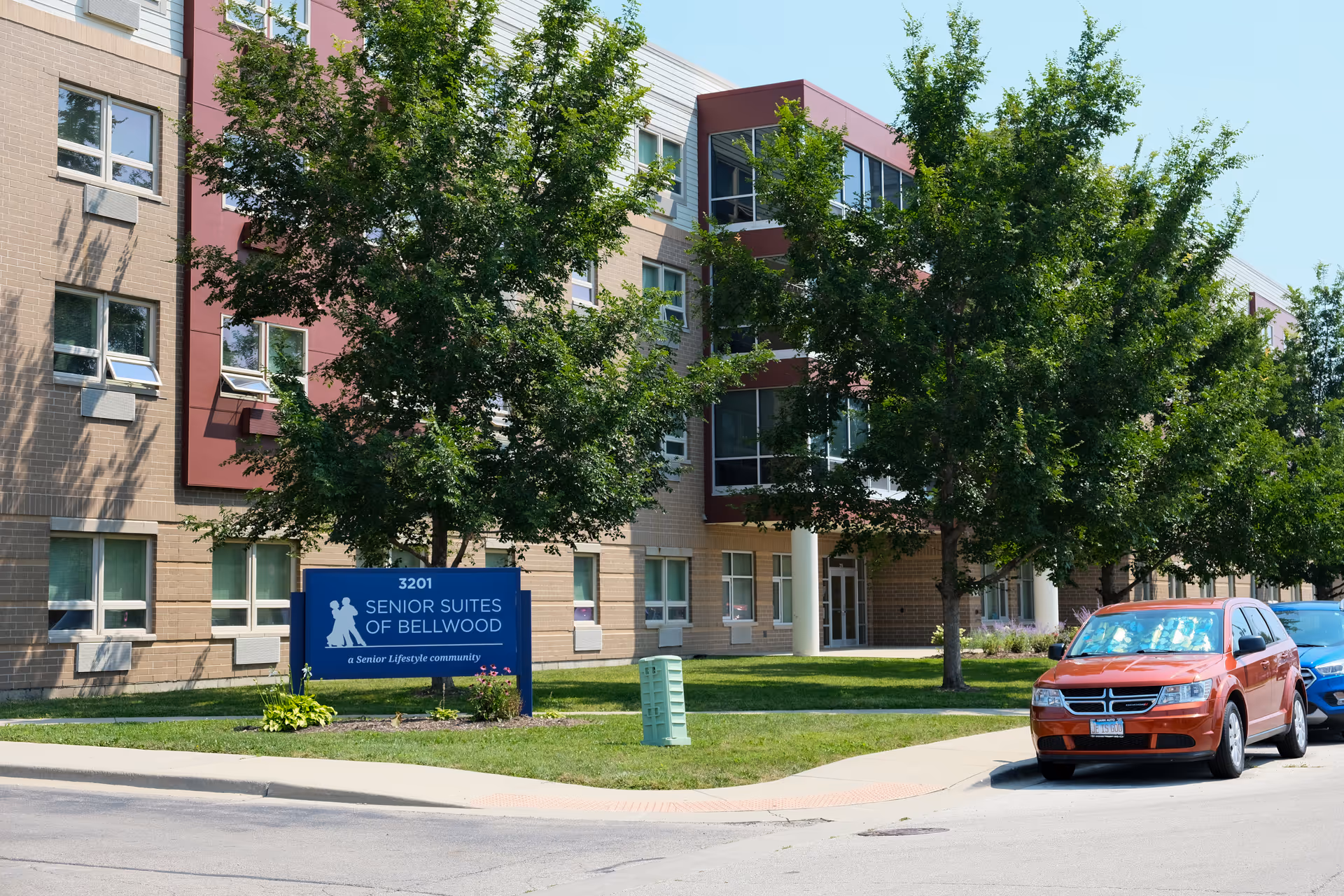Front exterior of a multi-story senior living building with a 'Senior Suites of Bellwood' sign, trees, and parked cars.