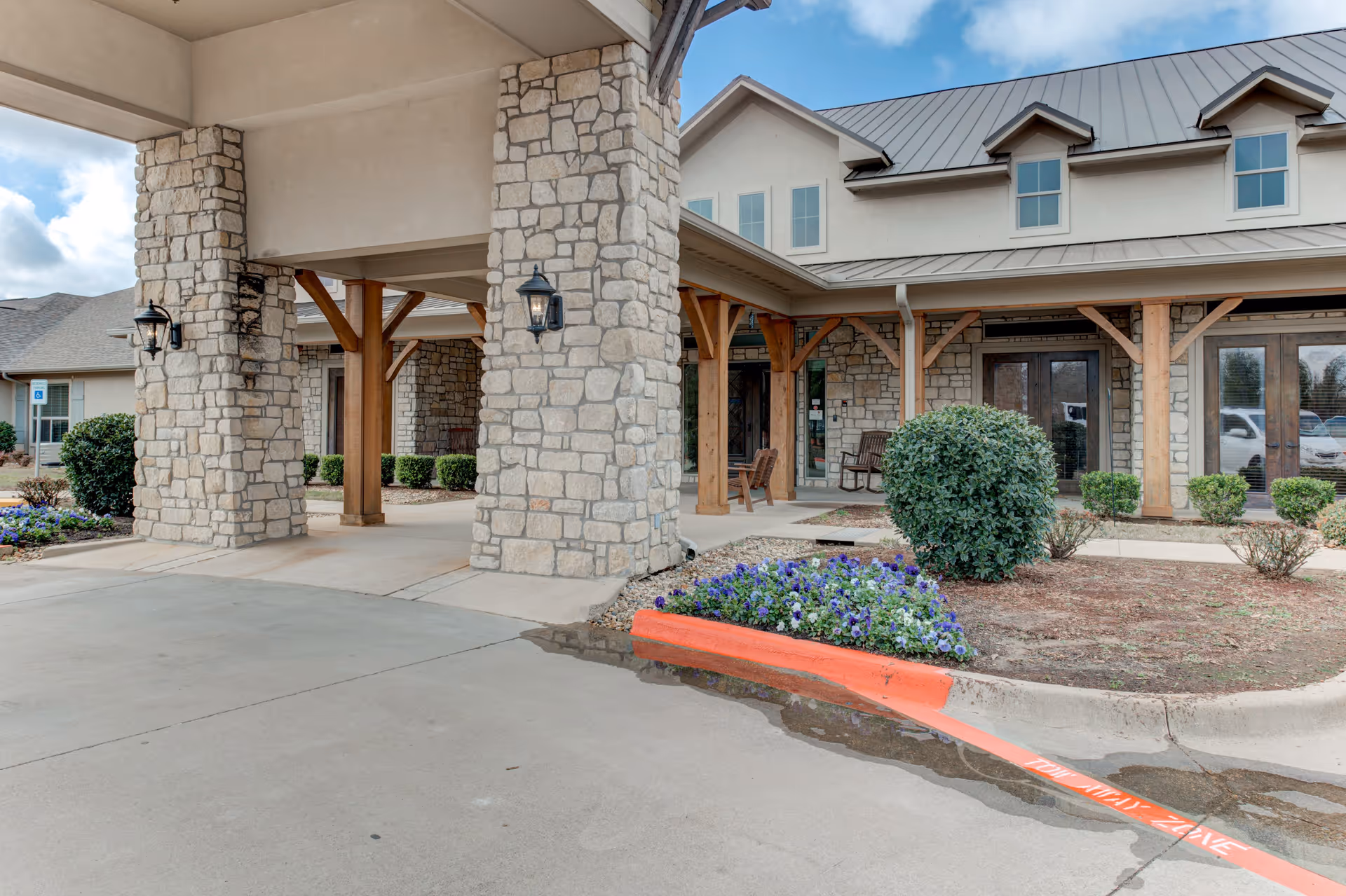 Entrance area of a senior living facility with stone pillars and wooden beams supporting a covered driveway. There are flower beds with purple flowers and green shrubs near the building, which has a stone facade and multiple windows. The sky is partly cloudy.
