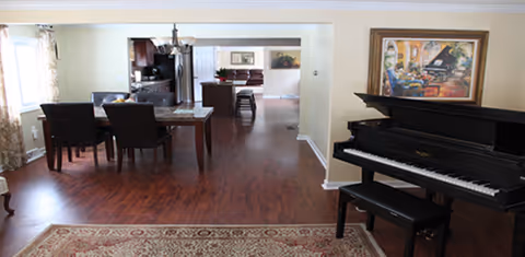 Open, well-lit senior living lounge featuring a piano, dining table, and dark wood flooring.