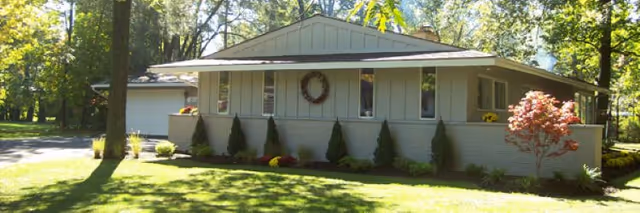 Single-story residential building with light siding, wreath, shrubs, and a lawn.