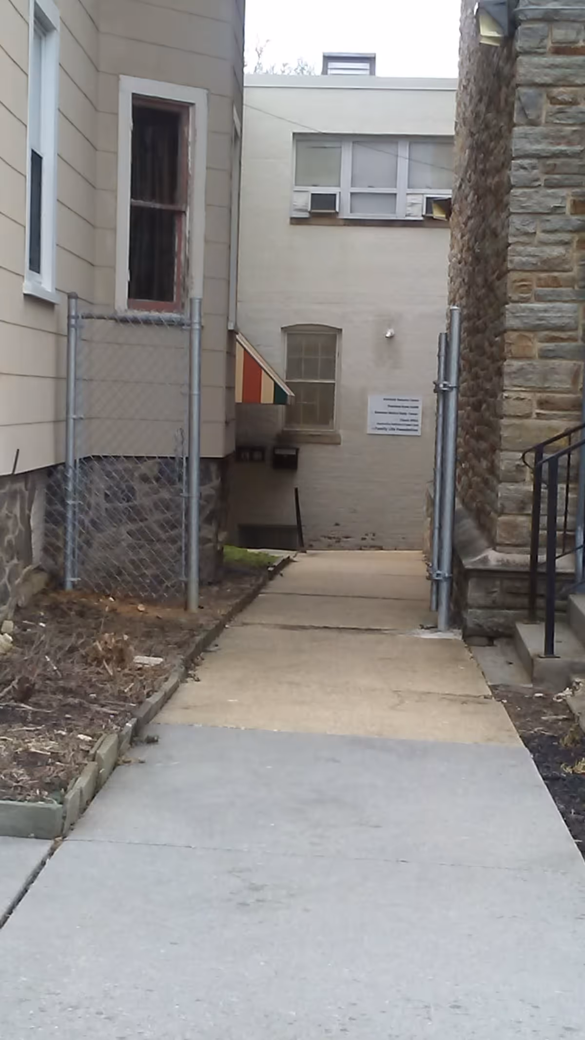Narrow concrete walkway between buildings with chain-link fencing and a stone wall.