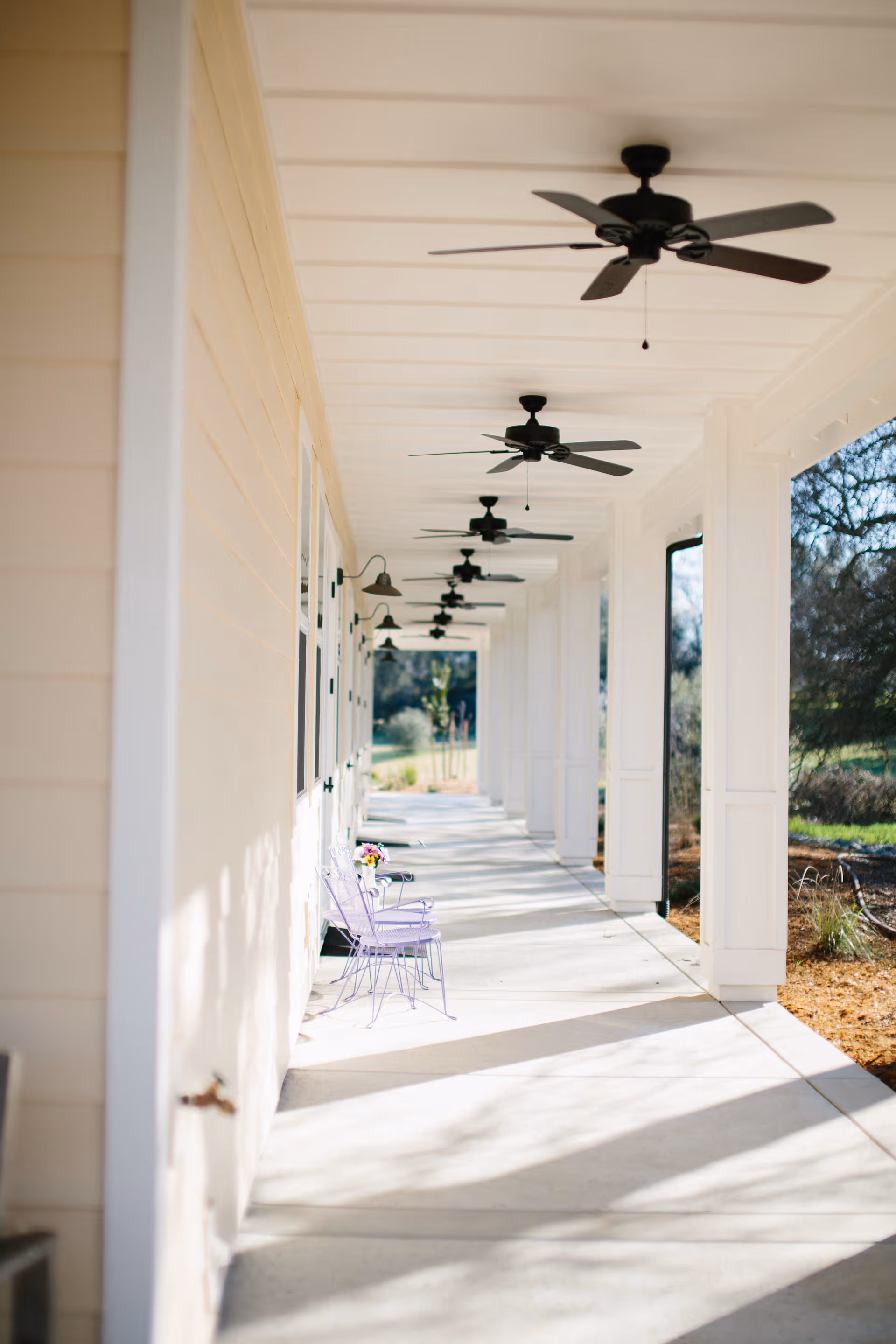 Long covered exterior porch with white columns, multiple ceiling fans, and a small table with pastel chairs along the walkway.