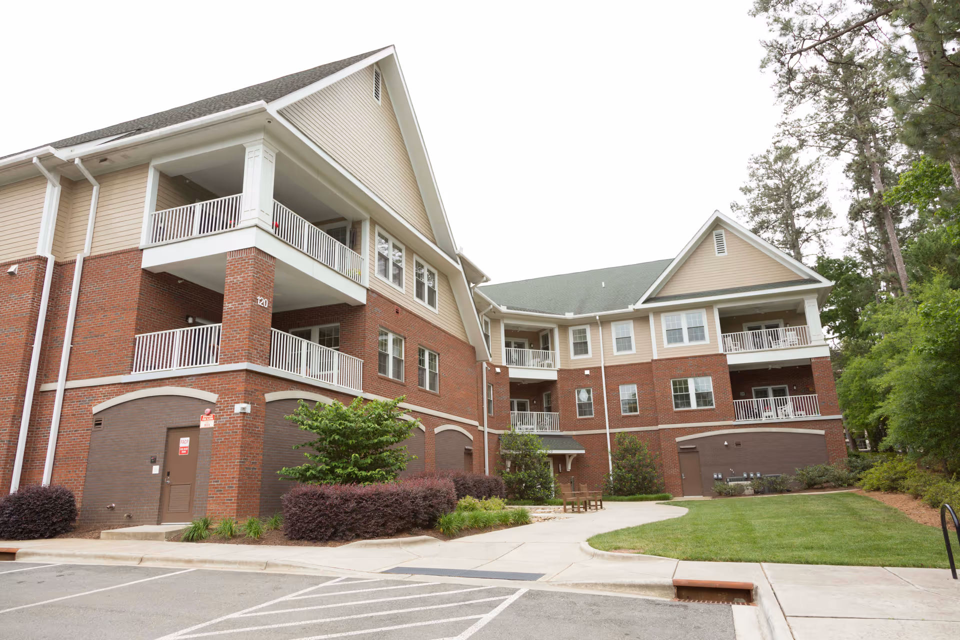 Exterior view of a multi-story senior living facility building with brick and beige siding, balconies, and surrounding greenery including trees and shrubs. There is a paved walkway and parking area in front of the building.