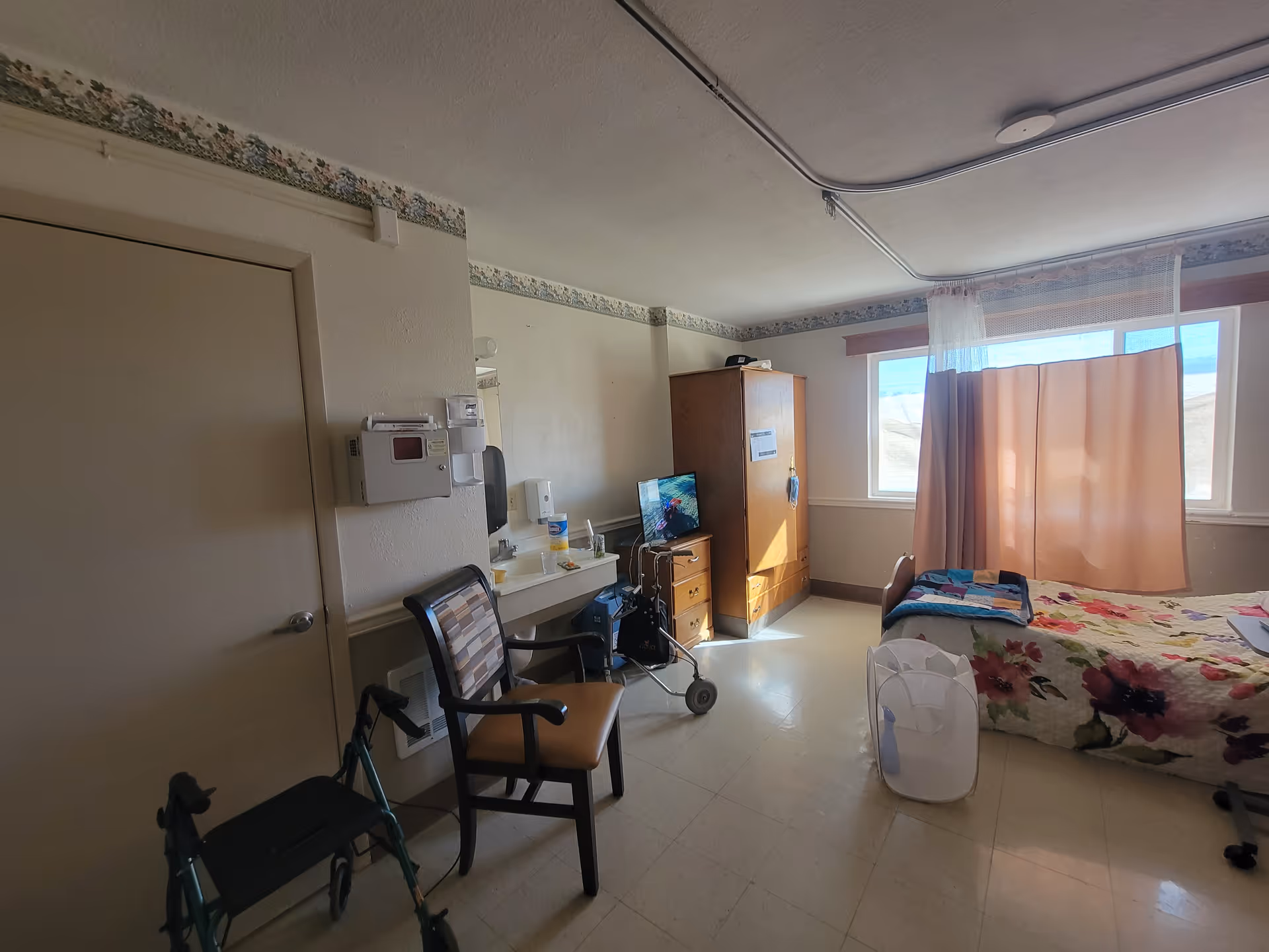 Interior senior living room with bed, wheelchair, walker, wardrobe, and pink-curtained window.