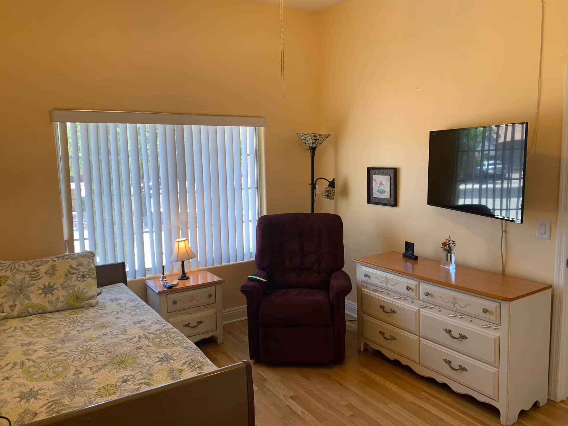 A cozy bedroom with bed, recliner, dresser, and mounted TV along pale yellow walls.