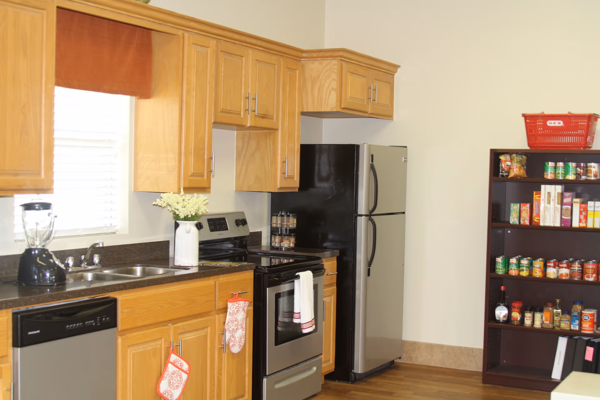 A kitchen area with wooden cabinets, a stainless steel refrigerator, stove, and dishwasher. There is a double sink under a window with blinds, a blender on the counter, and a vase with white flowers. On the right side, there is a shelving unit stocked with canned goods and other food items, with a red basket on top.