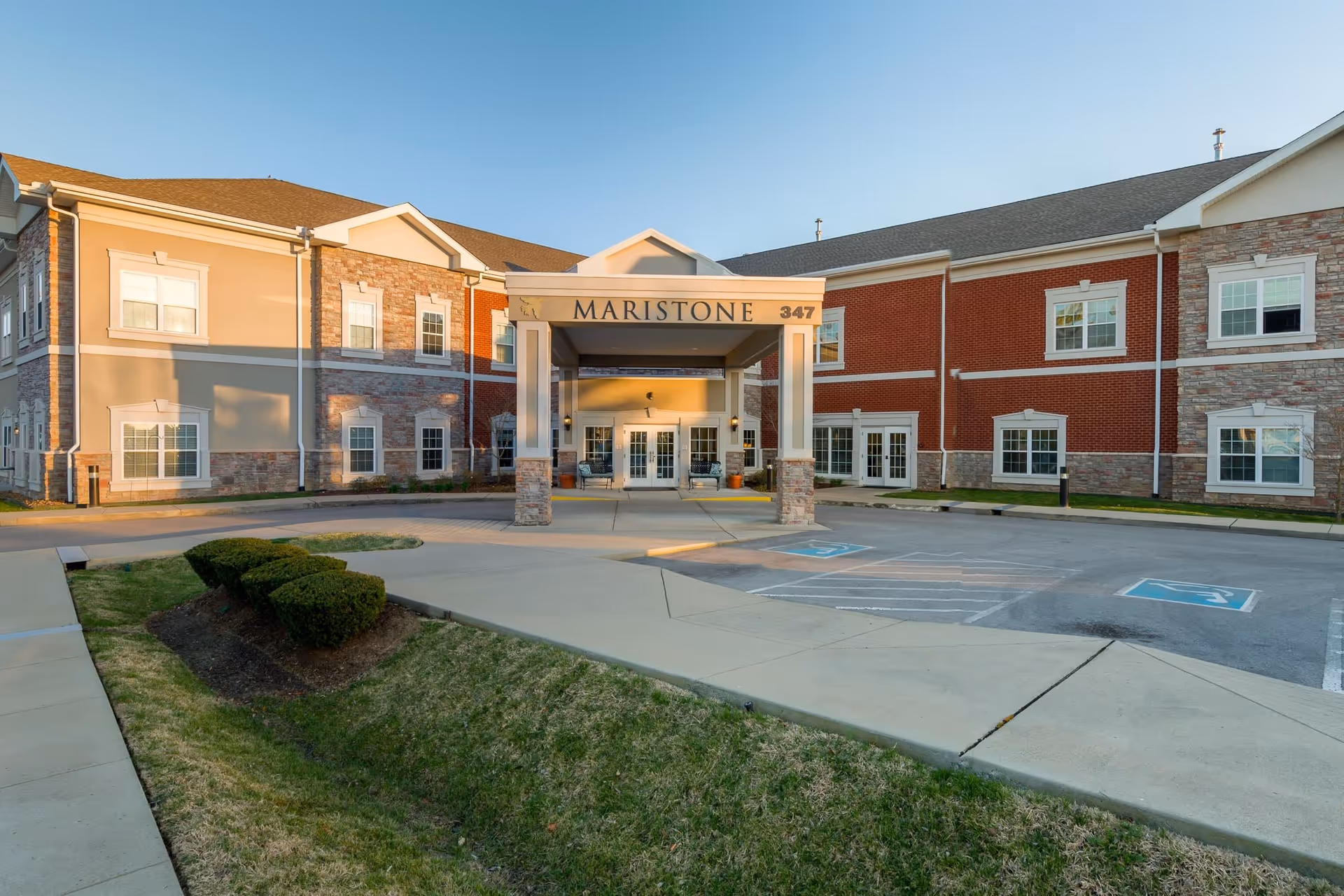 Exterior front view of Maristone of Franklin building with a covered entrance, multiple windows, and a parking area with handicap spaces. The building features a combination of brick and stone facade with a clear blue sky above.