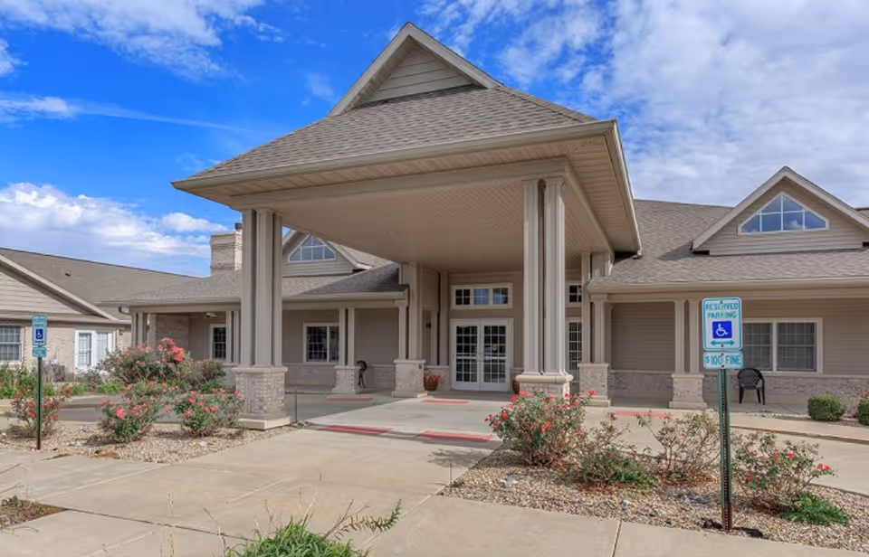 Front entrance of a senior living building with a covered porte-cochère, columns, landscaping, and handicap parking signs.