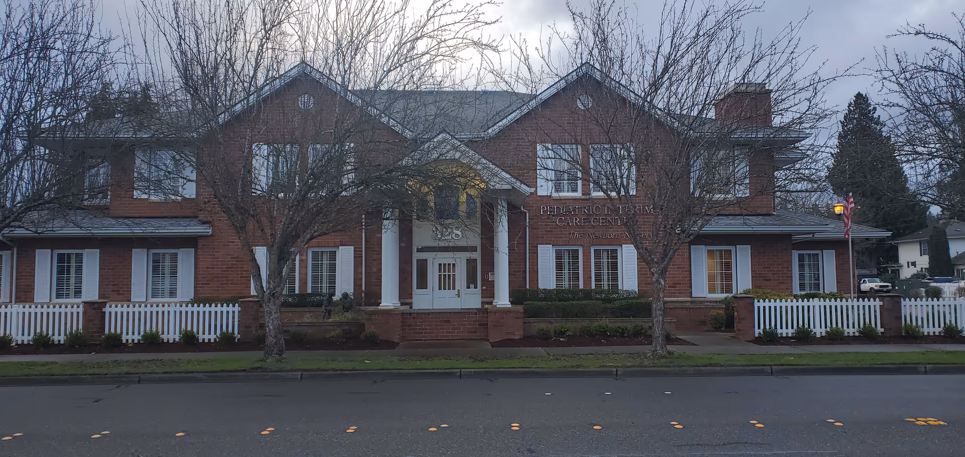 Brick facility with white porch, sign, and picket fence along a tree-lined front.