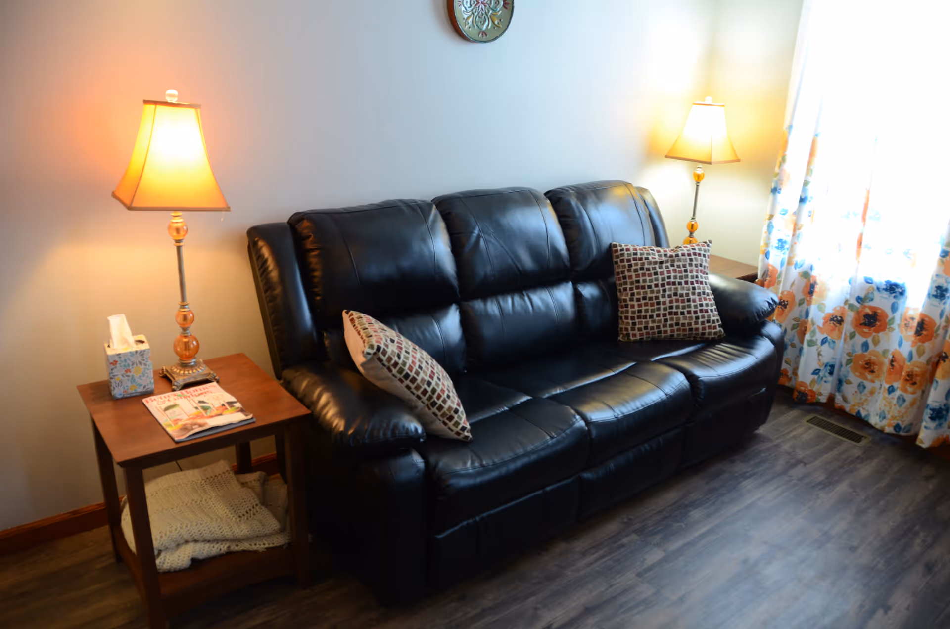 Cozy living room with black leather sofa, side tables, lamps, and floral curtains by a window.