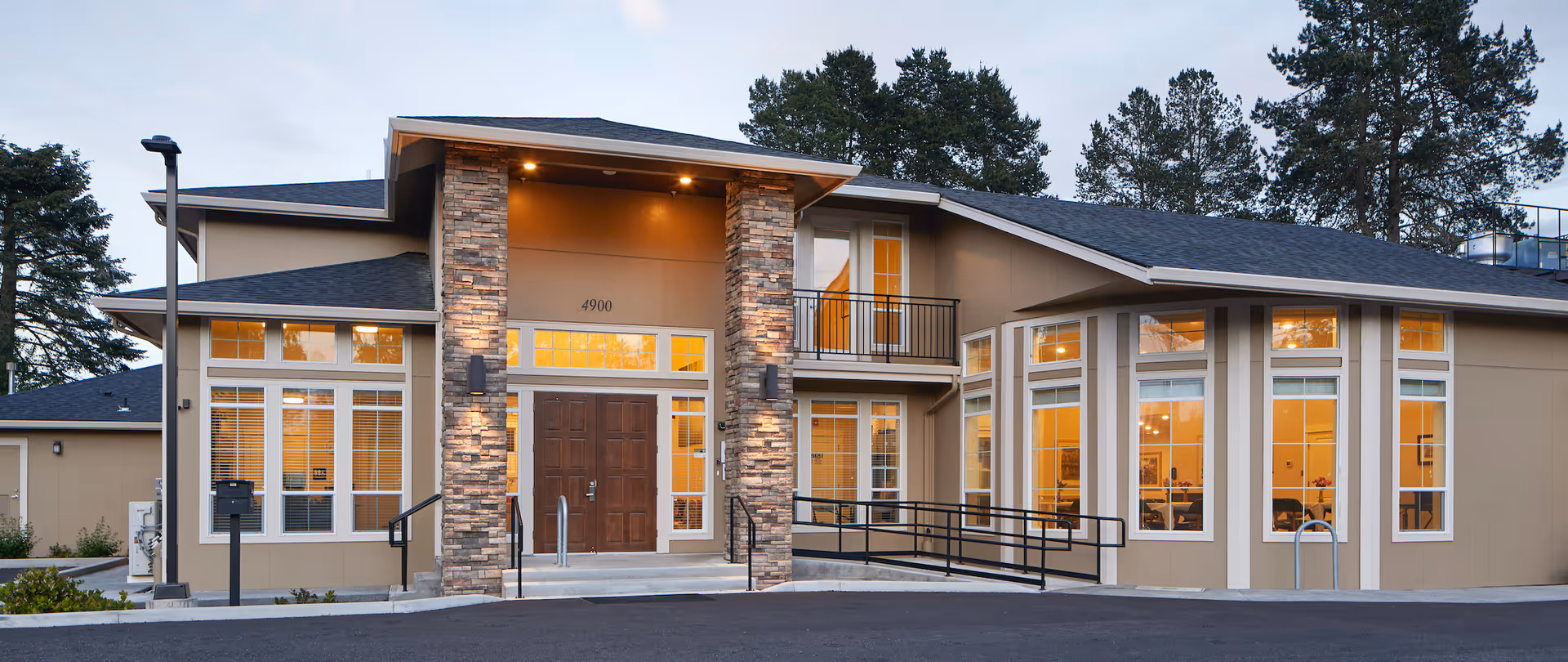 Exterior view of a modern senior living facility building with large windows, stone pillars flanking the entrance, a double wooden door, and a wheelchair ramp. The building is surrounded by trees and has warm interior lighting visible through the windows.