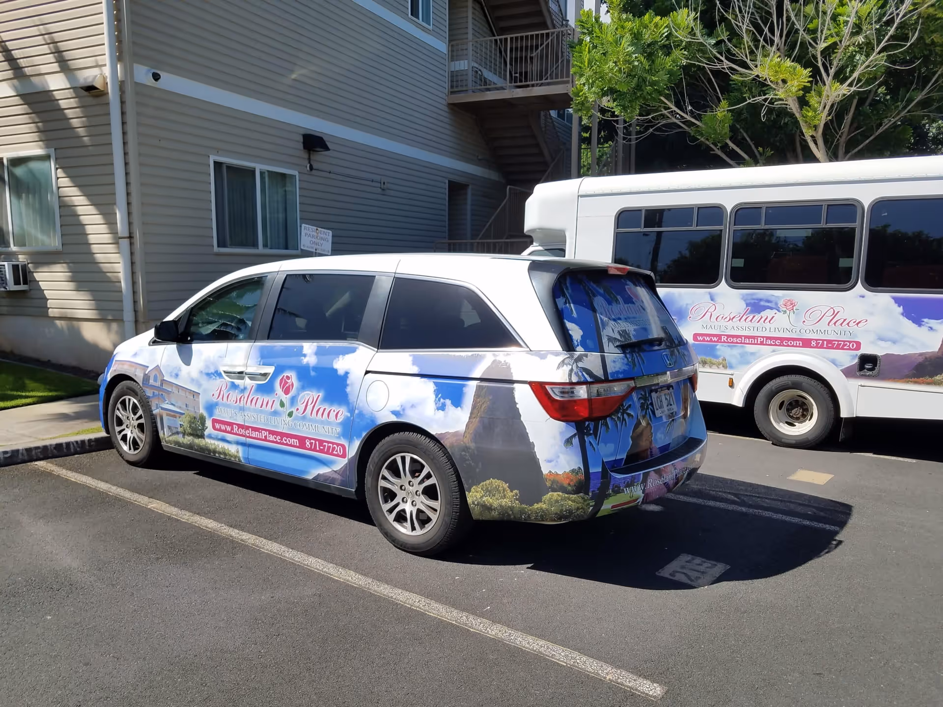 Two vehicles parked outside a building, both wrapped with advertisements for Roselani Place, an assisted living community. The vehicles display images of the facility, a rose logo, and contact information including a website and phone number.