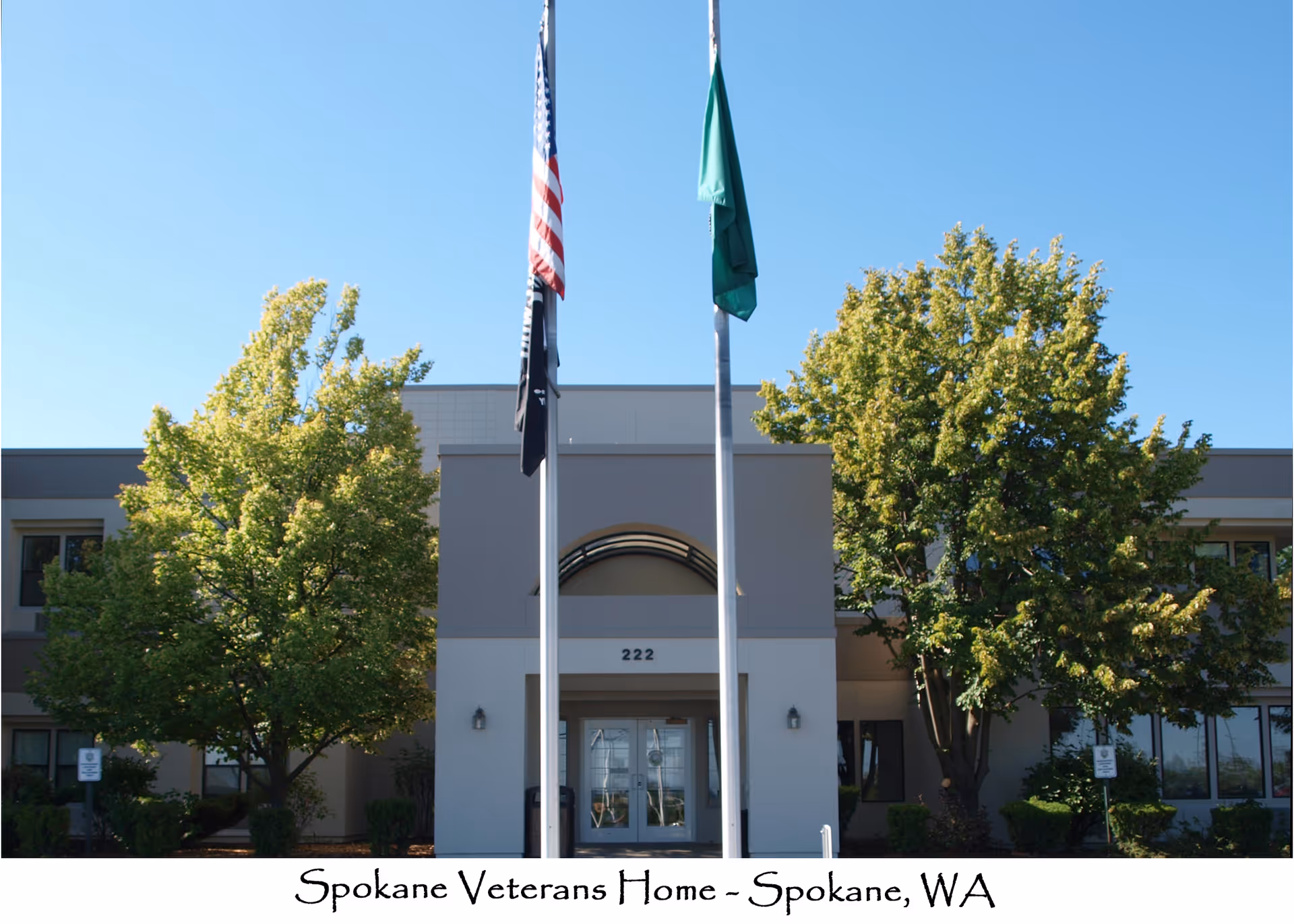 Front entrance of a two-story building with flagpoles and trees under a clear blue sky.
