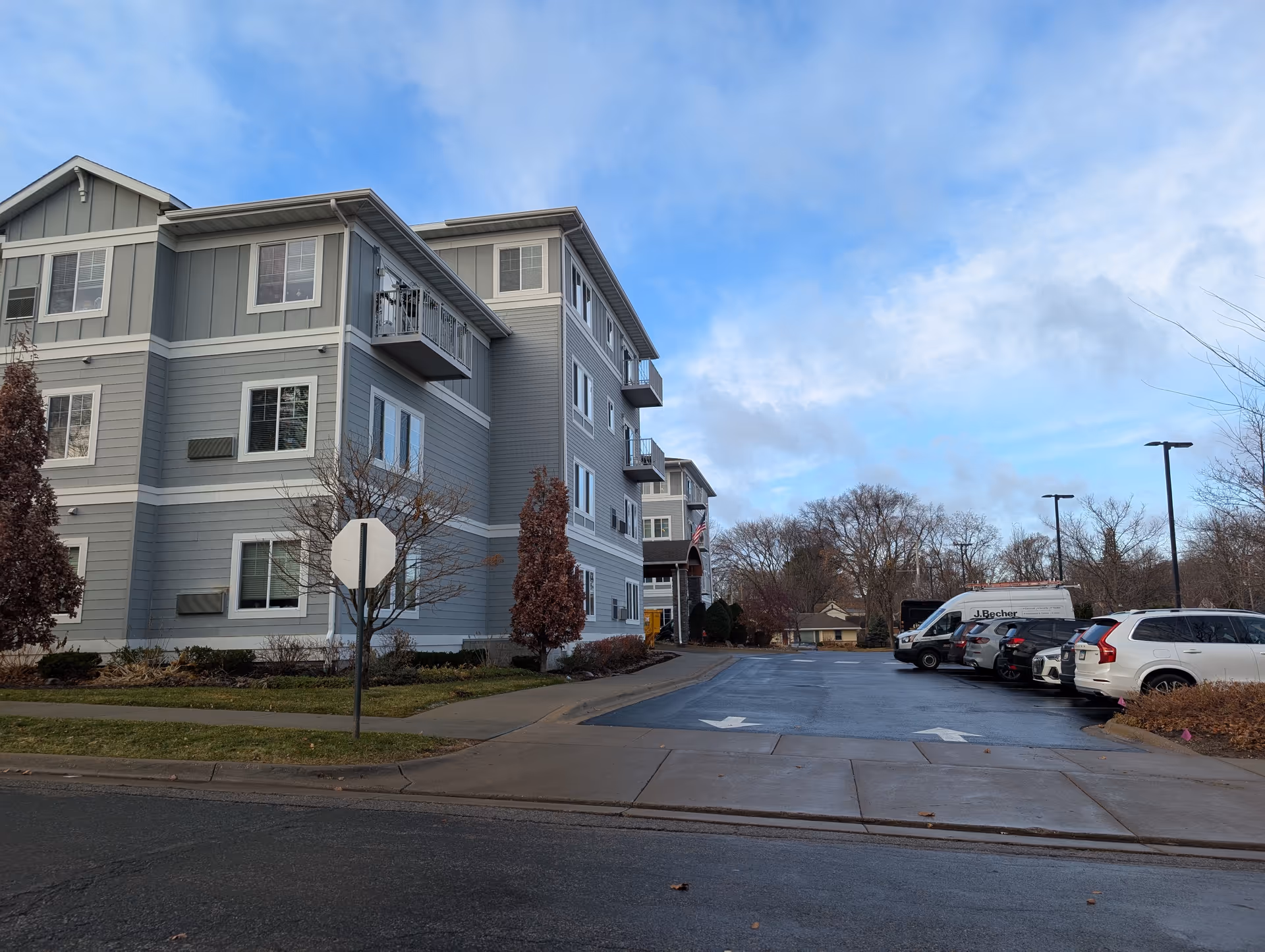 Front view of a gray multi-story senior living building with a driveway and parked cars under a blue sky.