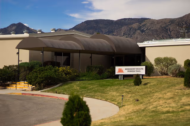Entrance to a single-story building with a covered walkway and a sign reading "Monument Health Pioneer Trail" set against a mountainous backdrop.