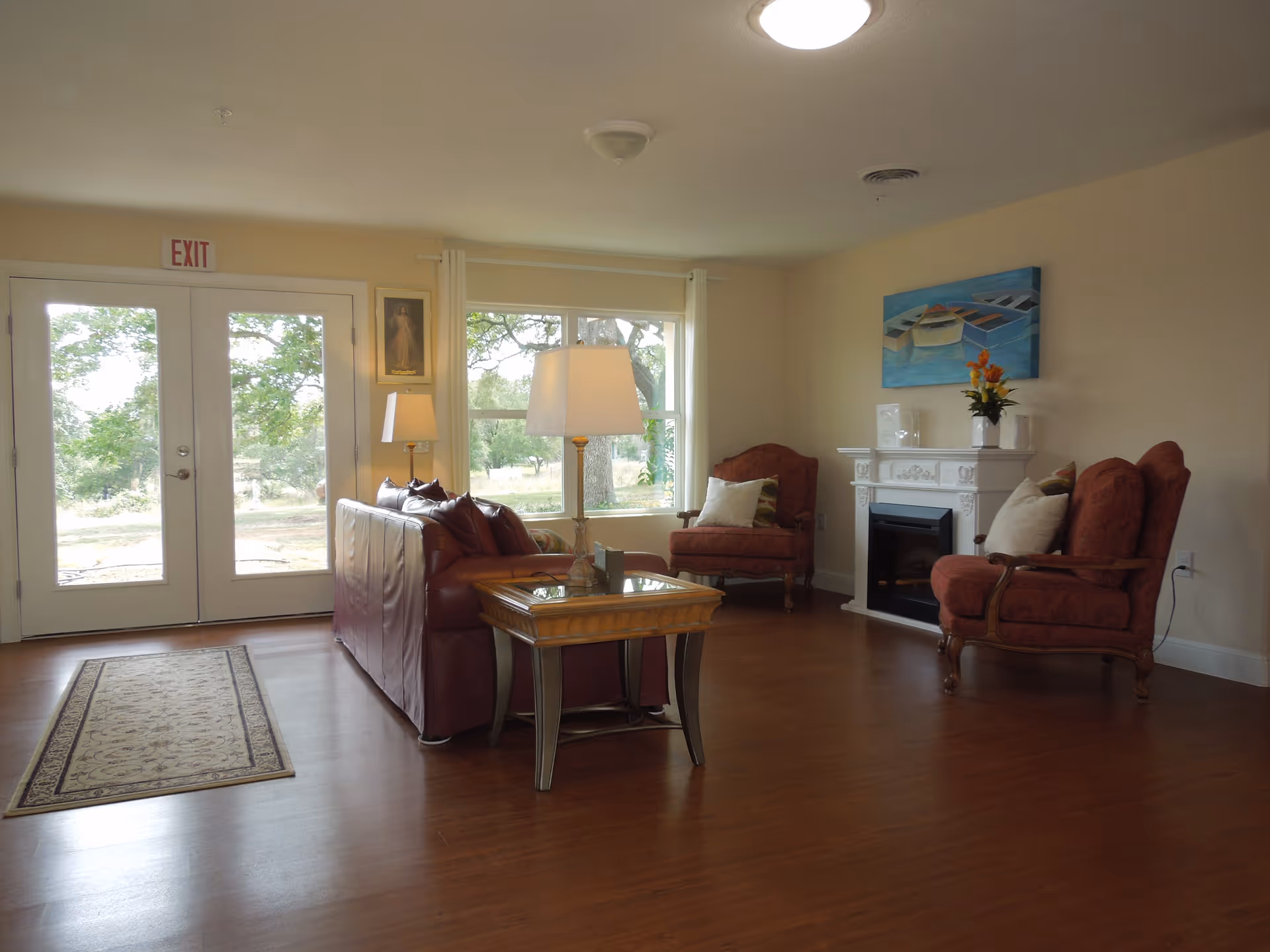 Bright communal living room with a leather sofa, upholstered armchairs, a coffee table, a fireplace, and large windows/doors.