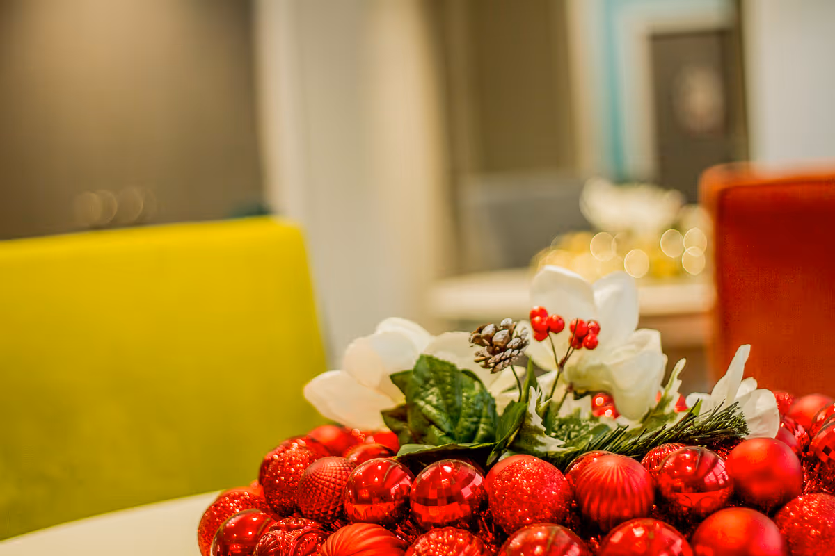 Close-up of a festive holiday centerpiece with red ornaments, white flowers, green leaves, and pinecones on a table, with blurred colorful chairs in the background.