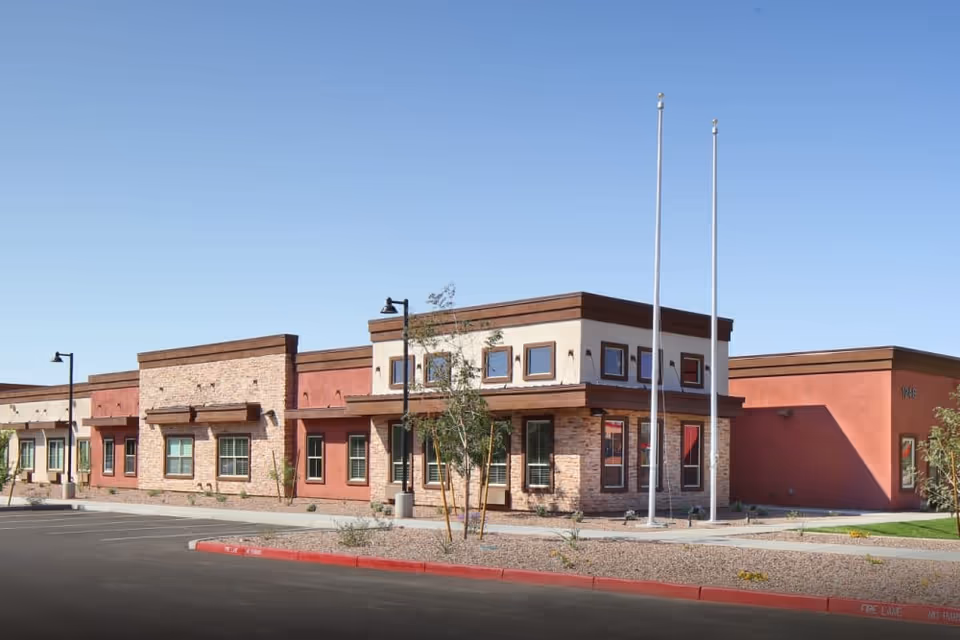 Exterior view of a single-story building with a modern southwestern architectural style, featuring a combination of stone and stucco walls, multiple windows, two flagpoles without flags, and a parking area in front under a clear blue sky.