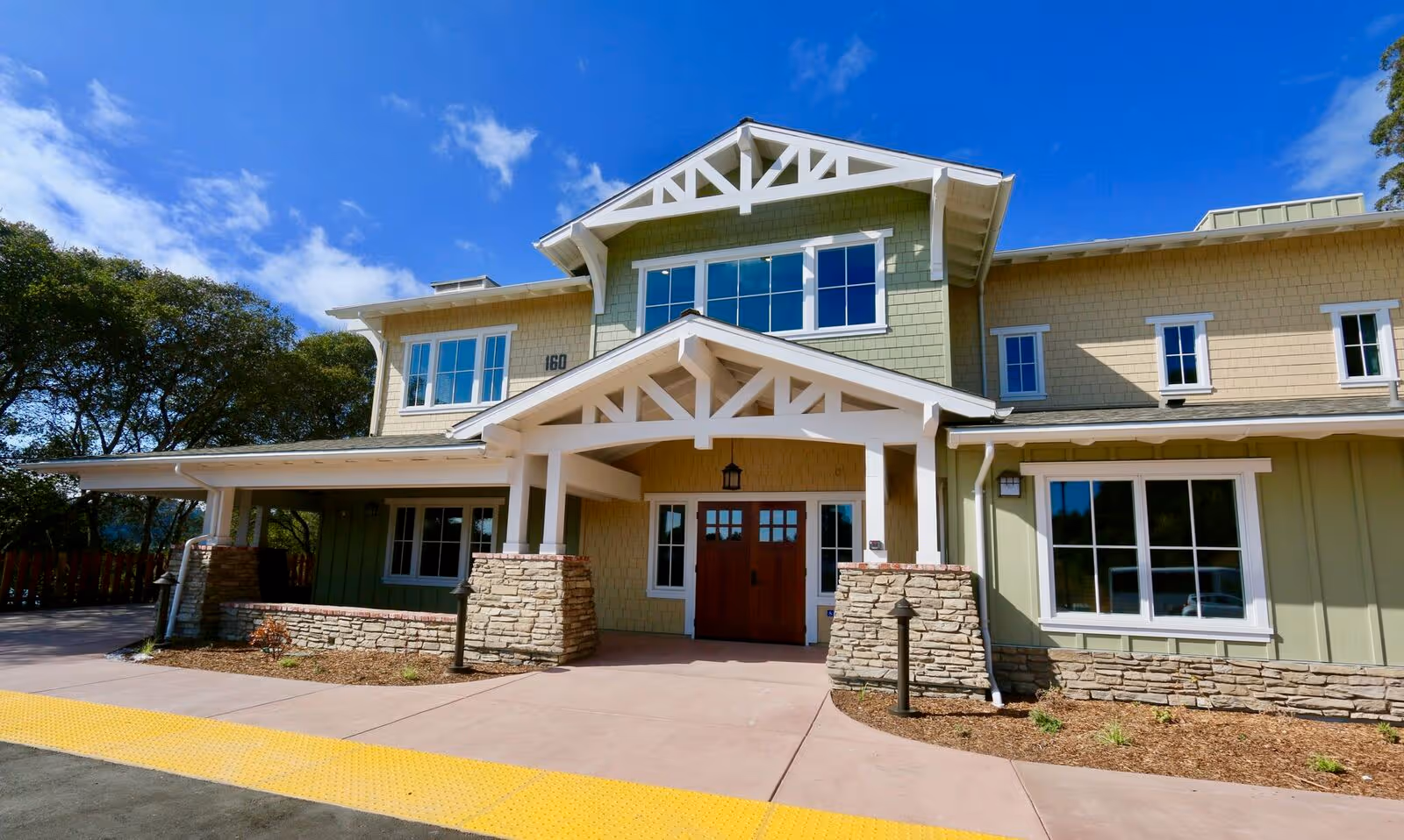 Exterior front view of a two-story building with green and beige siding, white trim, and a covered entrance supported by stone pillars. The building has multiple windows and a clear blue sky with some clouds in the background.