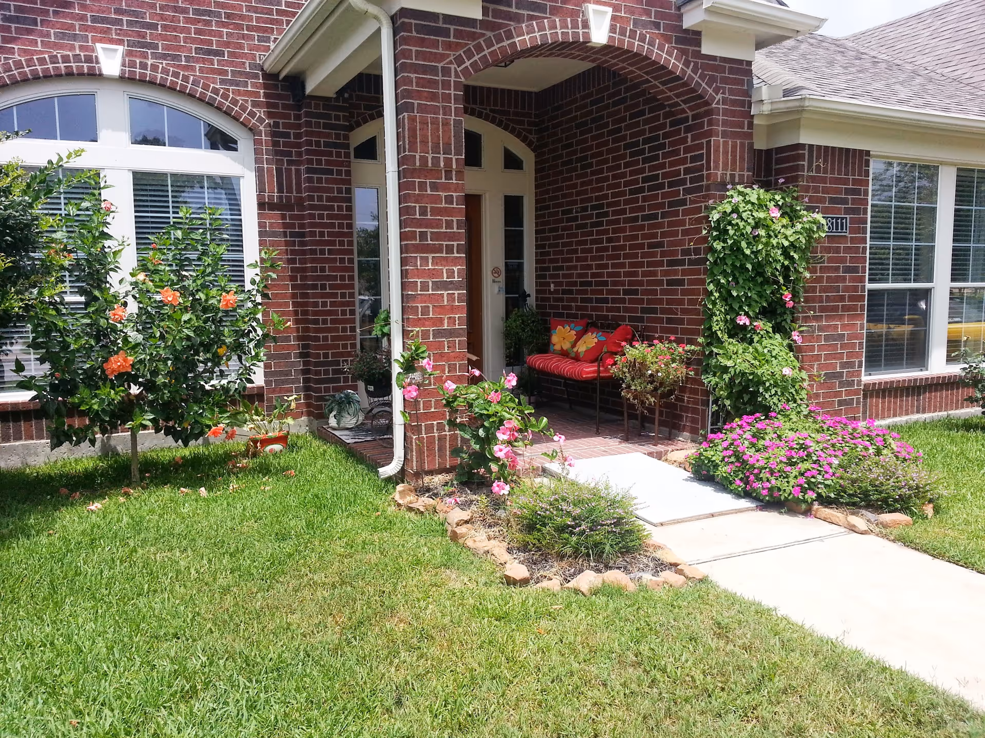 Brick-front senior living facility entrance with a flower-filled garden and porch seating.