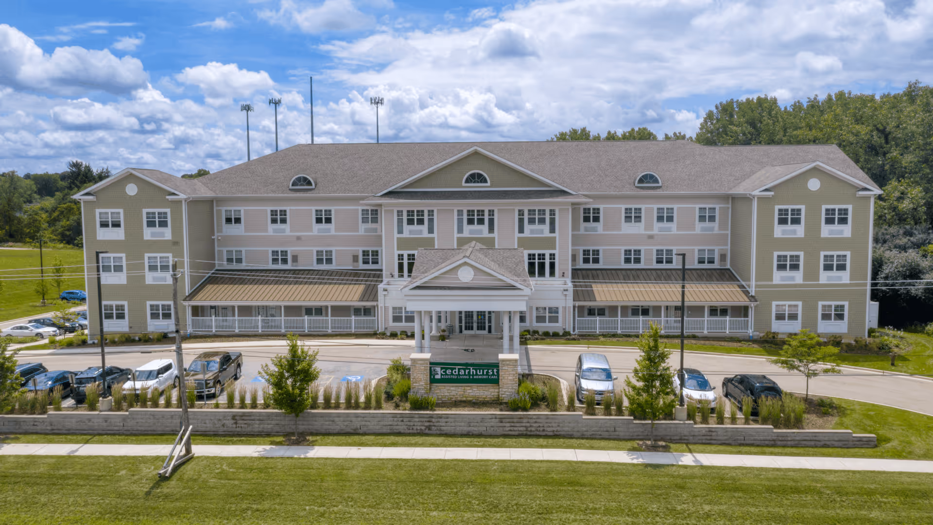 Front exterior view of Cedarhurst Senior Living of Naperville, a three-story building with multiple windows, a covered entrance, and a parking area with several cars. The building is surrounded by green lawns and trees under a partly cloudy sky.