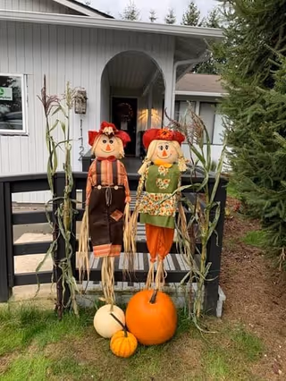 Front entrance of a light-gray house decorated with two scarecrows, pumpkins, and cornstalks on the porch railing.