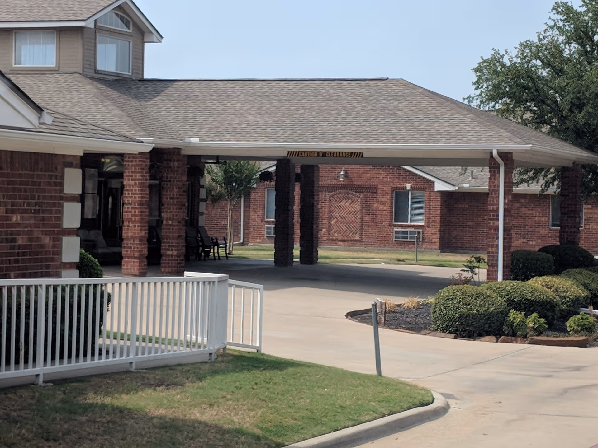 Brick senior living facility entrance under a covered driveway, with shrubs and white railing.