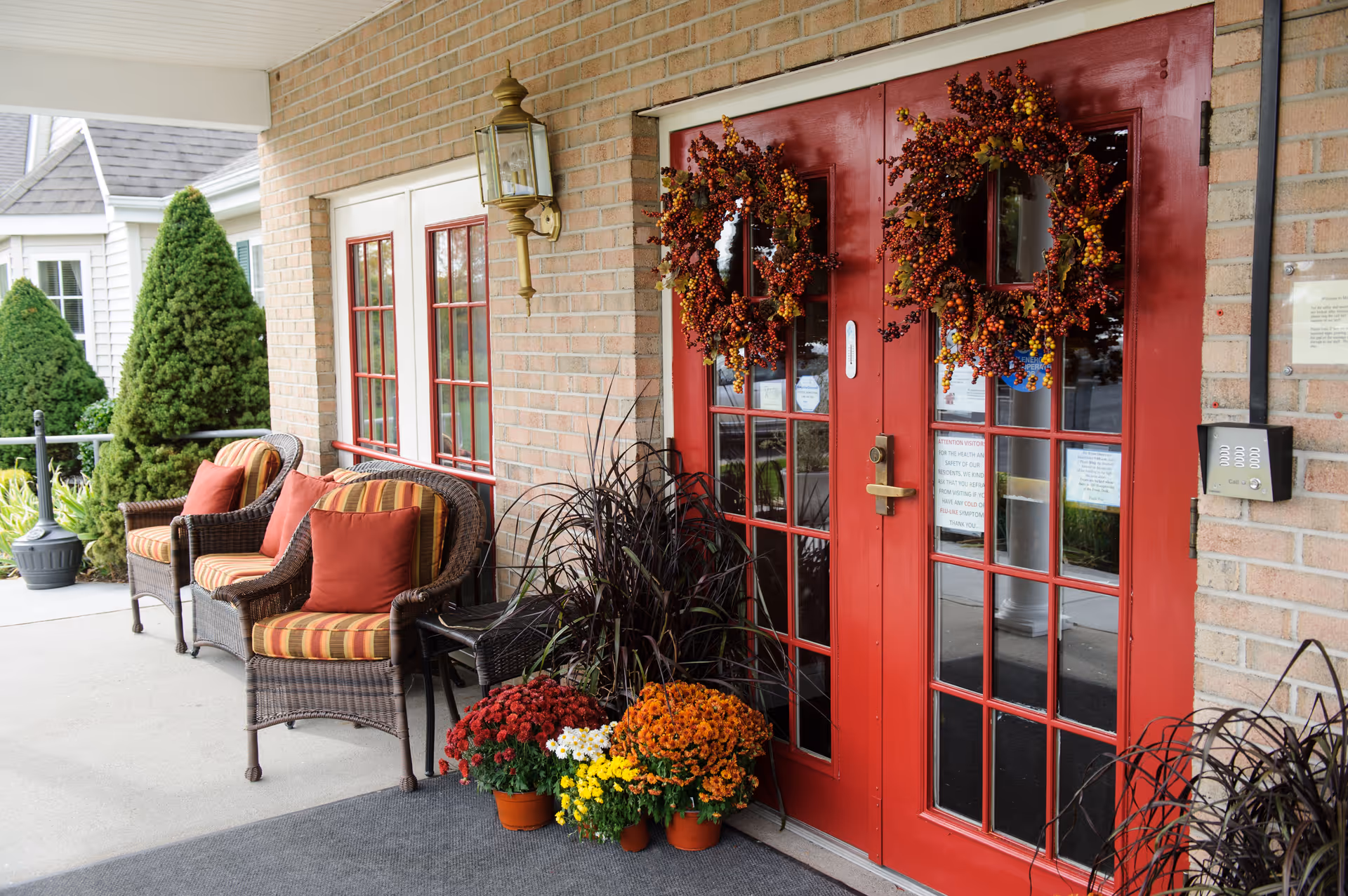 Entrance to a facility with double red doors decorated with autumn wreaths. There are wicker chairs with striped cushions and red pillows on the left side, along with potted flowers and plants near the door. The building exterior is made of light-colored bricks, and there is a lantern-style light fixture mounted on the wall.