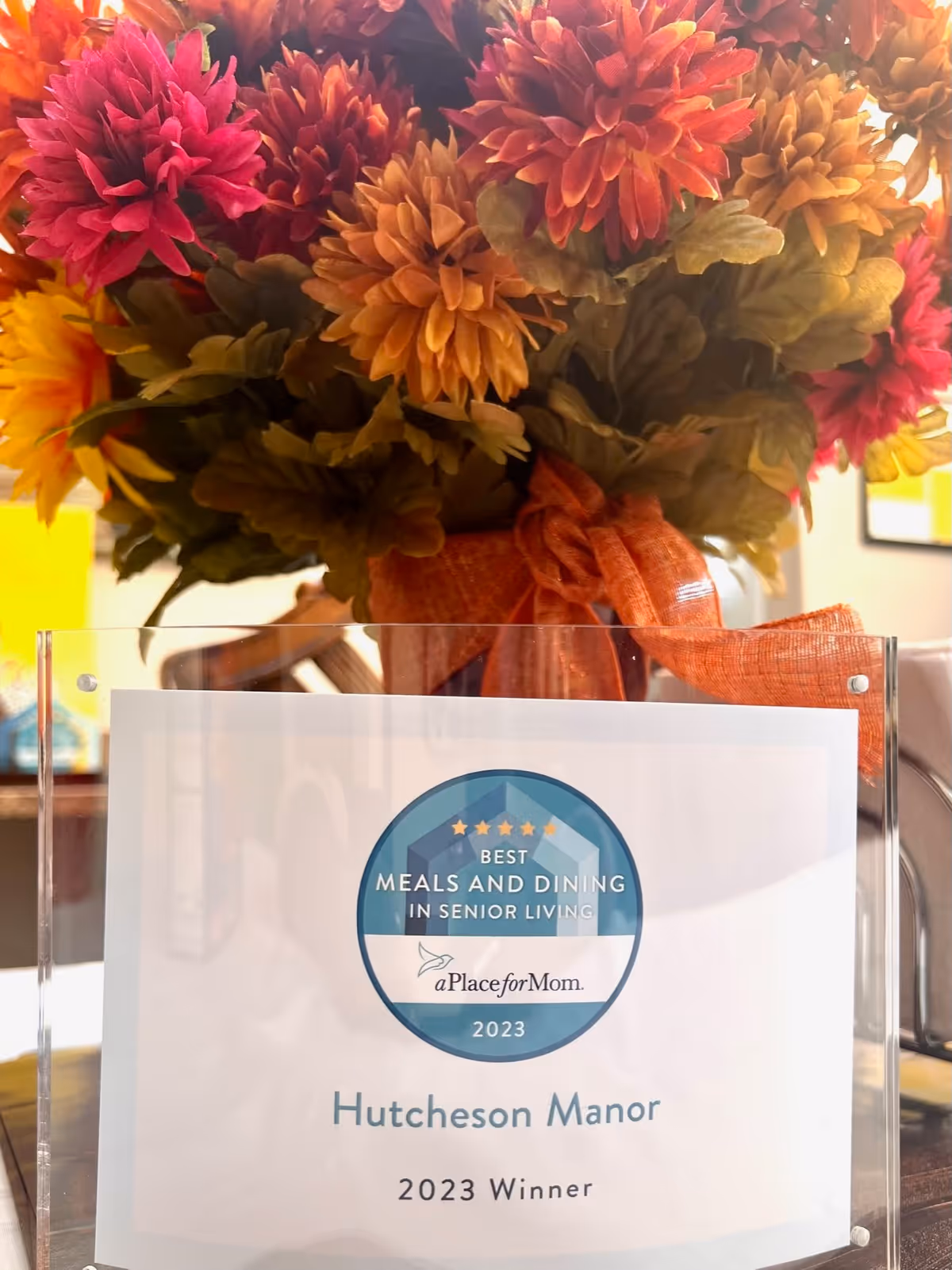 A close-up image of a floral arrangement with orange, red, and yellow flowers tied with an orange ribbon. In front of the flowers is a clear plaque that reads 'Best Meals and Dining in Senior Living, aPlaceforMom, 2023, Hutcheson Manor, 2023 Winner.'