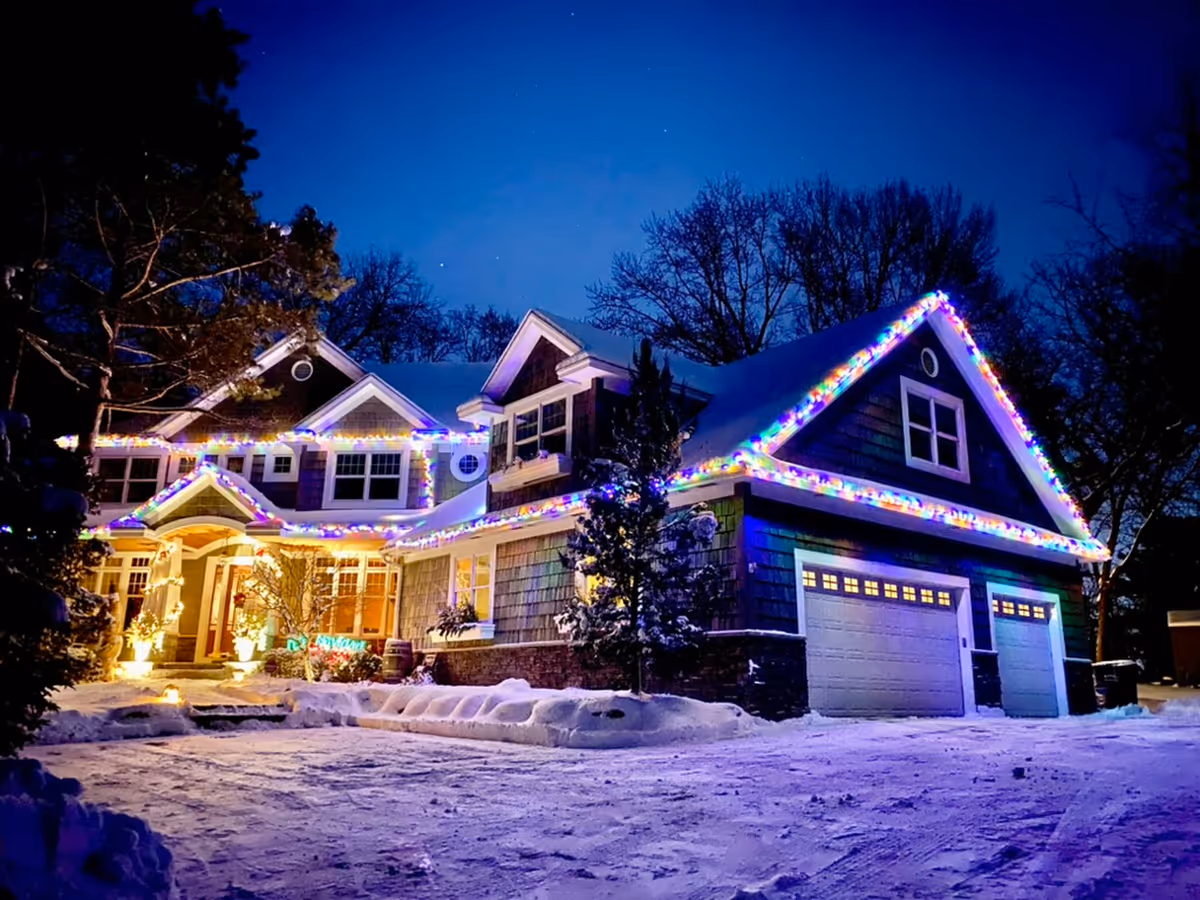 Snowy night exterior of a house with colorful Christmas lights along the rooflines.