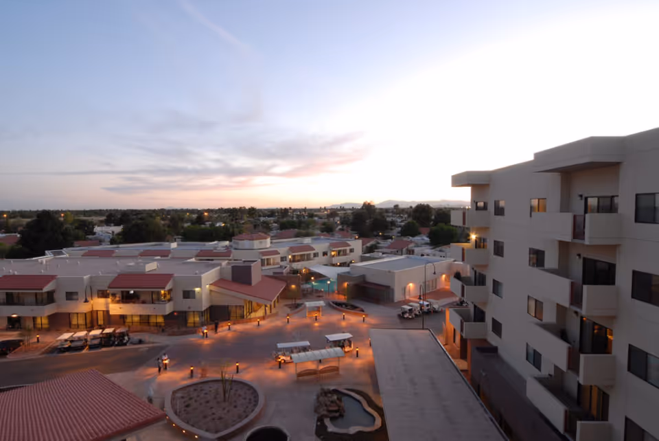 Aerial view of Friendship Village Of Tempe senior living facility at dusk, showing multiple buildings with balconies, outdoor seating areas, pathways, and landscaped spaces under a partly cloudy sky.