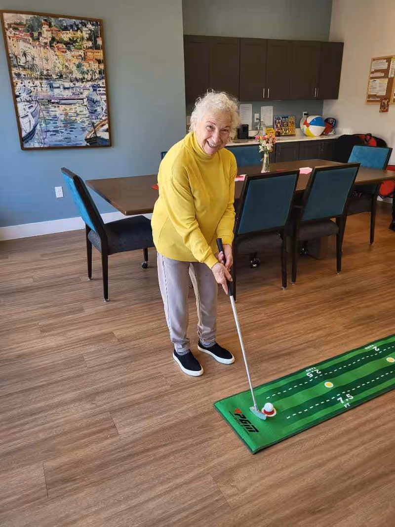 An elderly woman wearing a yellow sweater and gray pants is playing indoor mini golf on a green putting mat inside a room with wooden flooring. Behind her is a long table with blue chairs, a vase with flowers, cabinets, and a colorful painting on the wall.