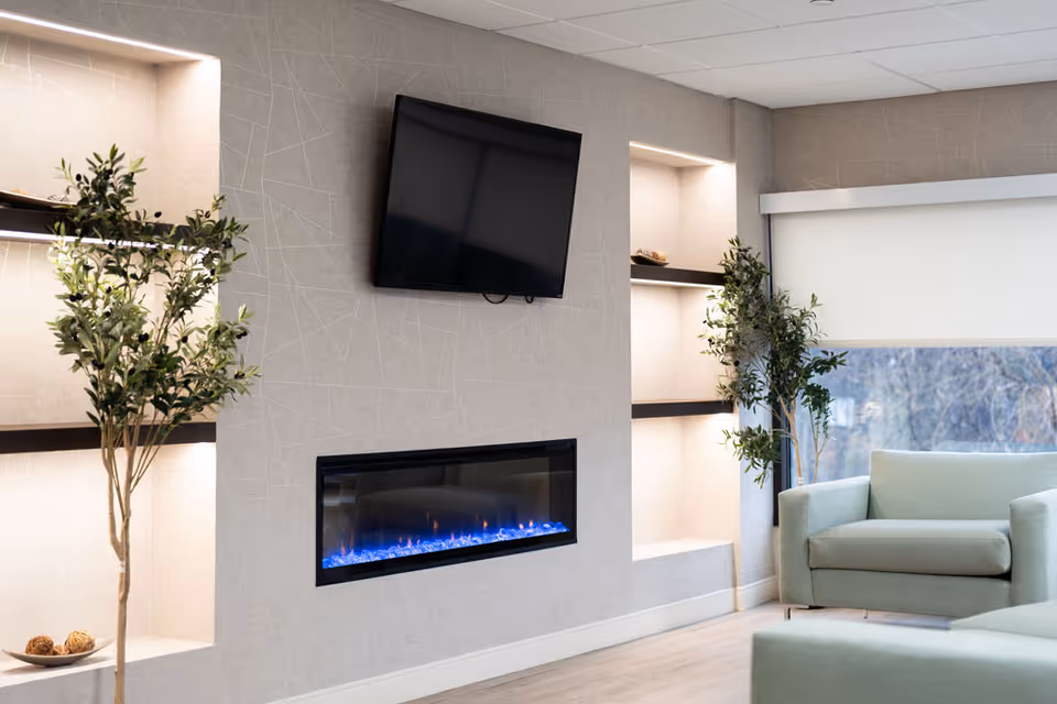 Modern senior living lounge with fireplace, wall-mounted TV, illuminated shelves, and green sofas.