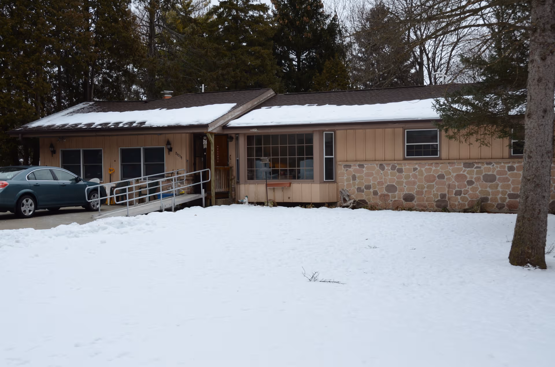 Single-story beige building with stone facade and snow-covered yard, parked car beside ramp.