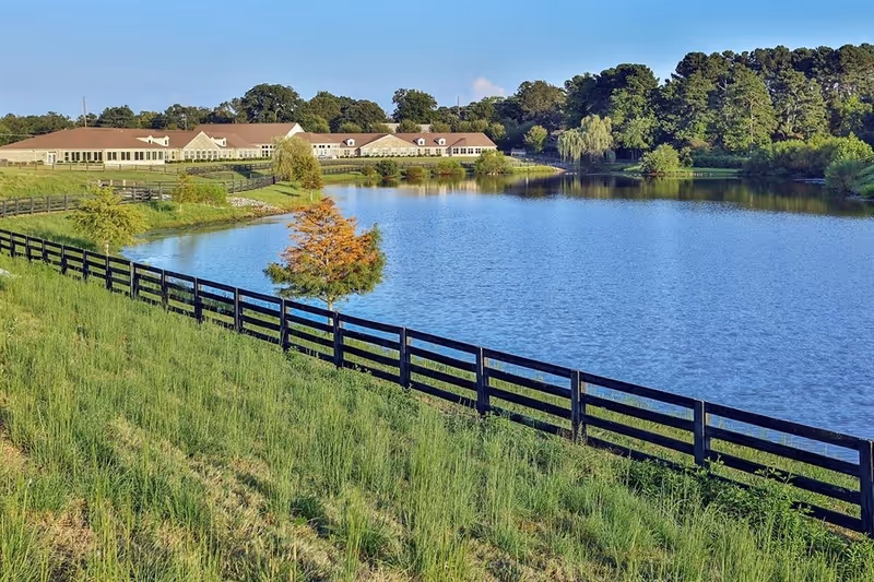 A senior living campus sits beside a calm lake, with a wooden fence and green fields.