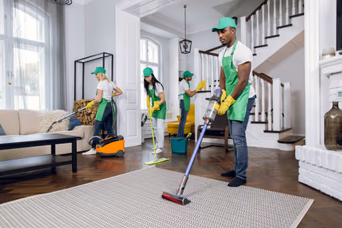 Four cleaning staff in green aprons clean a bright living room.