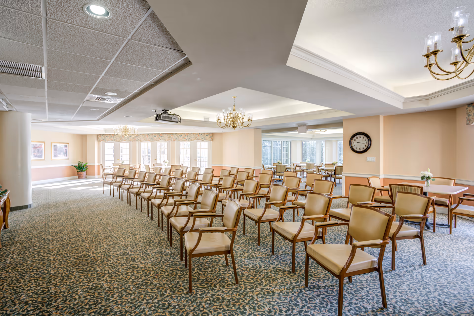 Large conference room with rows of beige chairs, chandeliers, and windows inside a senior living facility.
