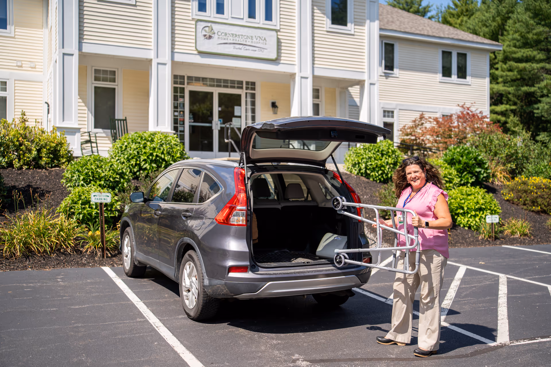 A woman with a walker stands beside an open SUV trunk at Cornerstone VNA facility entrance.