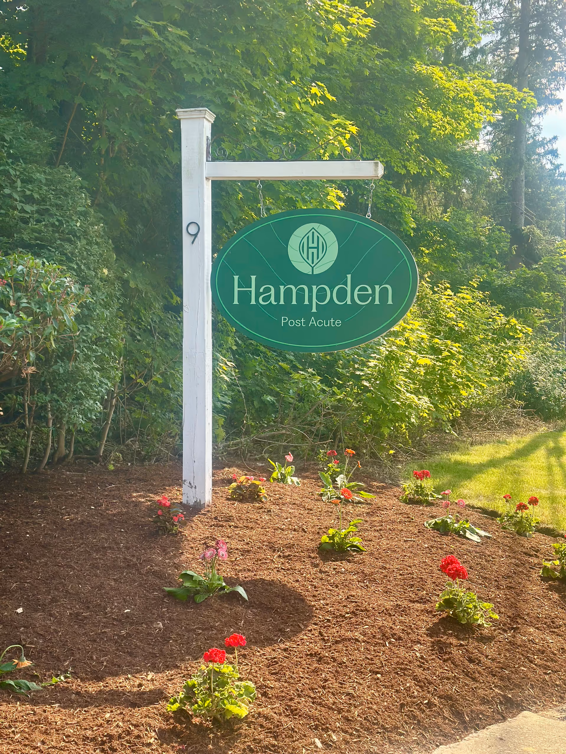 Green oval sign reading Hampden Post Acute hangs from a white post beside flowers.