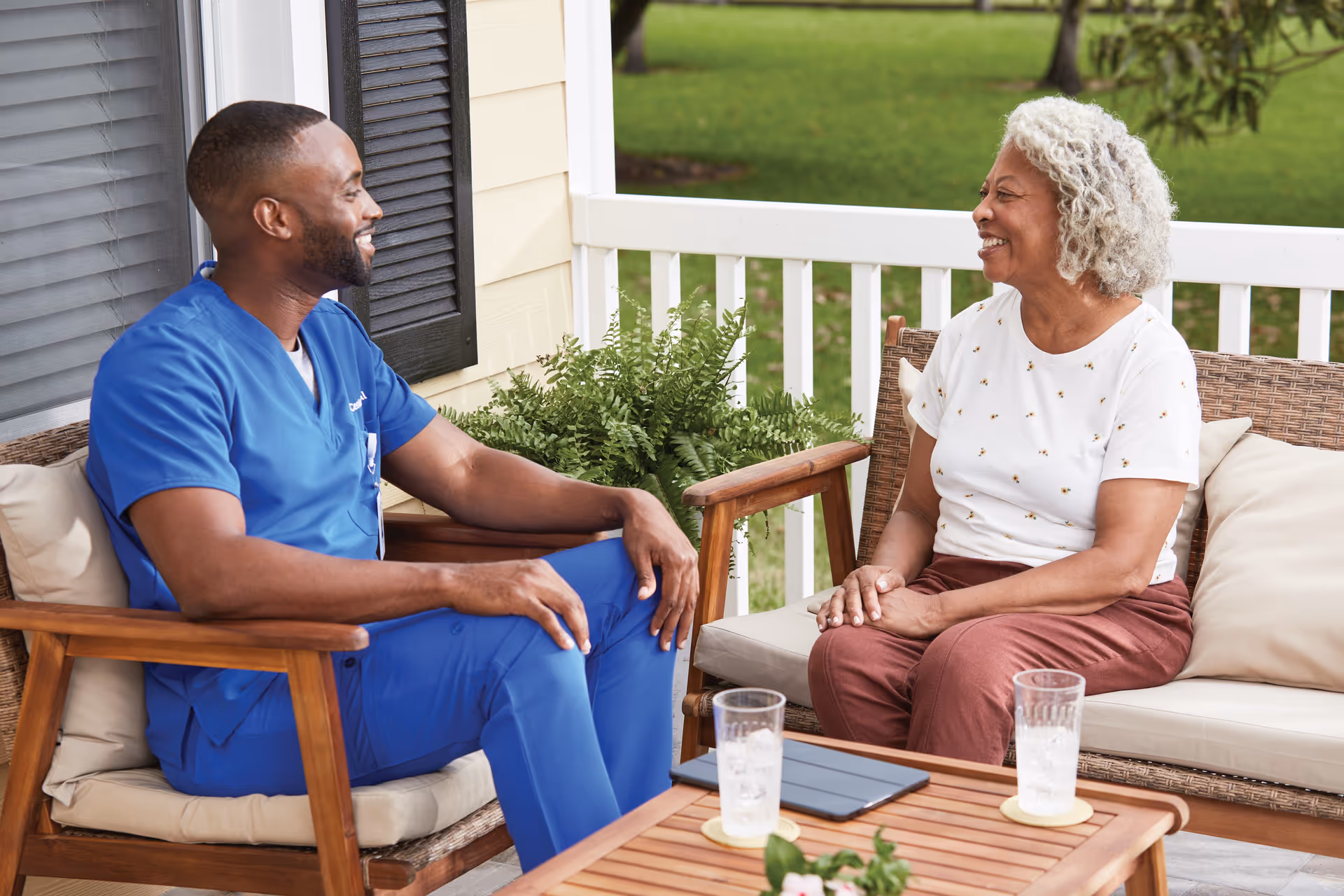 Healthcare worker in blue scrubs chats with an elderly woman on a sunny outdoor porch.