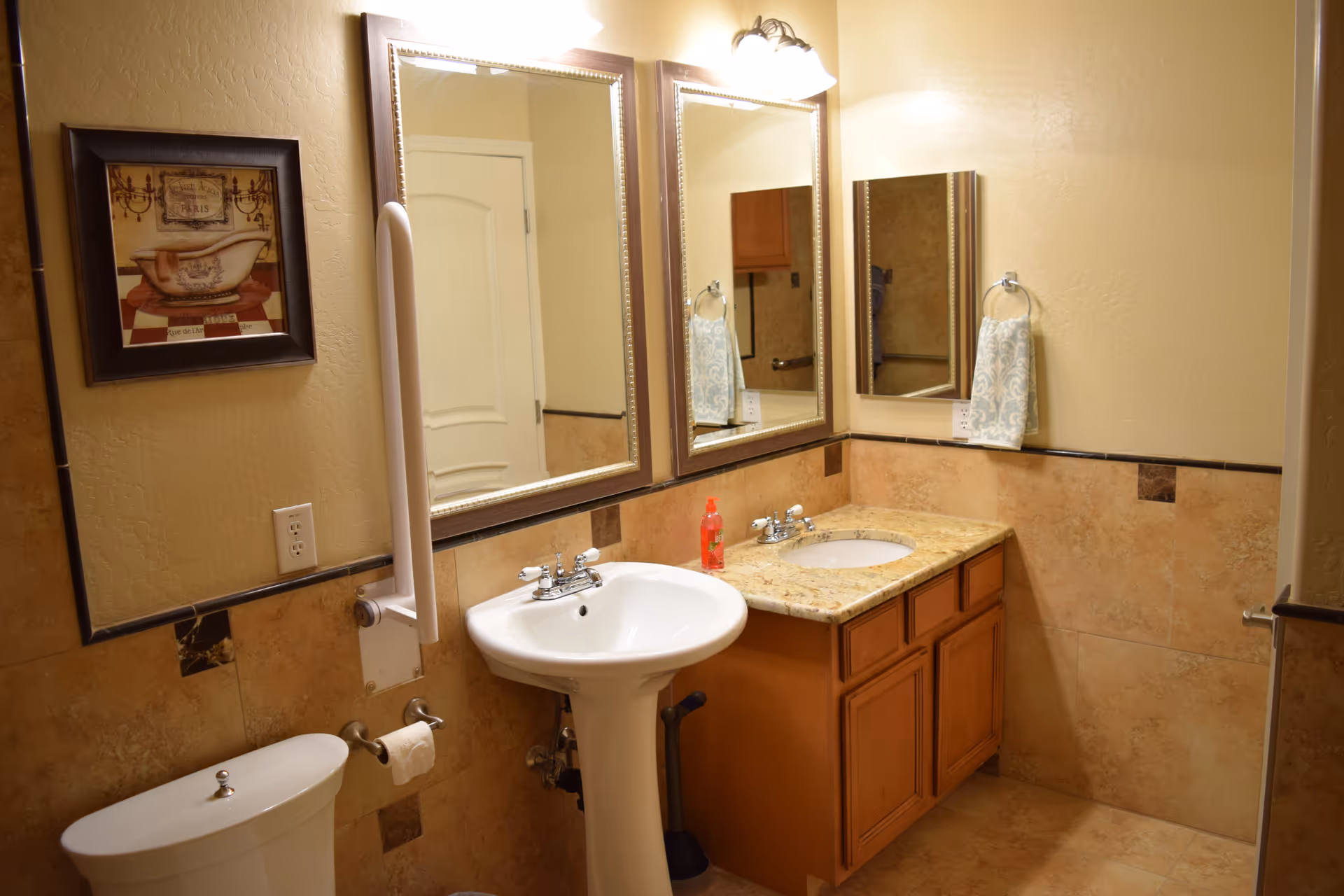 A bathroom with beige tiled walls and floor, featuring a white toilet, a pedestal sink, and a vanity with a granite countertop and built-in sink. Two large framed mirrors are mounted above the sinks, with a light fixture above them. A hand towel hangs on a ring next to the vanity, and a framed picture of a bathtub is on the wall above the toilet. A grab bar is installed next to the toilet for support.