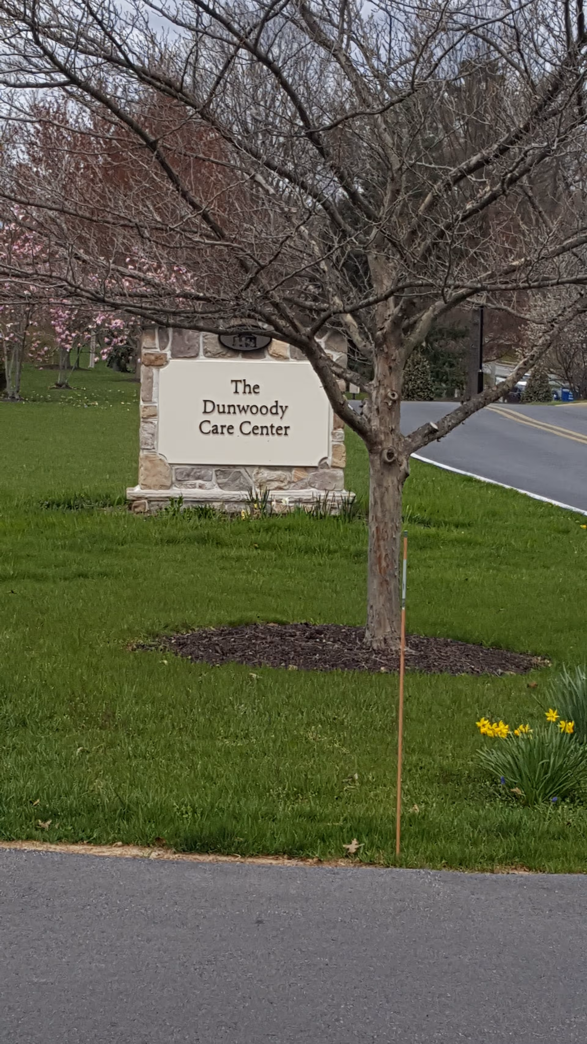 A stone sign reads The Dunwoody Care Center beside a tree and grassy lawn.