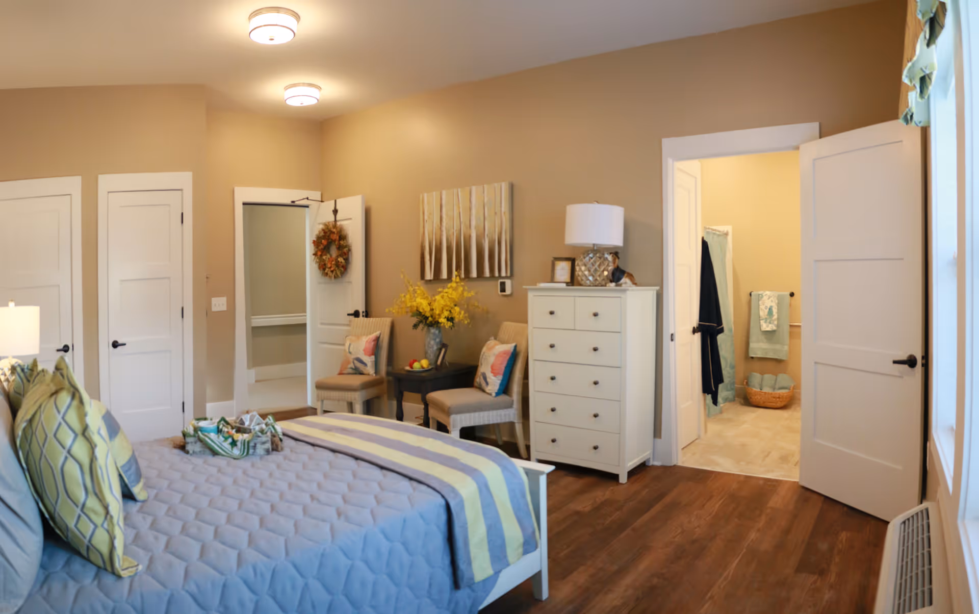 Furnished bedroom with a made bed, dresser, chairs and an open door leading to a bathroom.