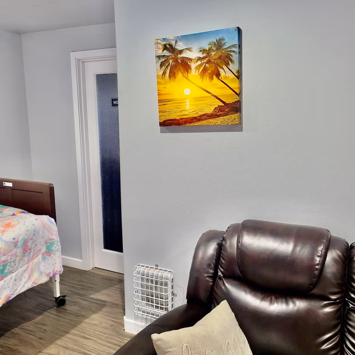 A cozy bedroom corner with a bed, brown recliner, vent heater, and palm artwork.