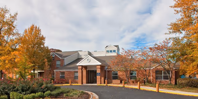 Exterior view of a single-story brick building with a peaked roof and a covered entrance. The building is surrounded by trees with autumn foliage and a curved driveway leading to the entrance under a partly cloudy sky.