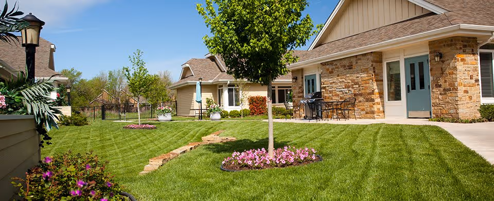 A well-maintained outdoor garden area at Veranda Senior Living featuring green grass with striped mowing patterns, a small tree surrounded by pink flowers, stone edging, patio furniture, and buildings with stone and beige siding under a clear blue sky.