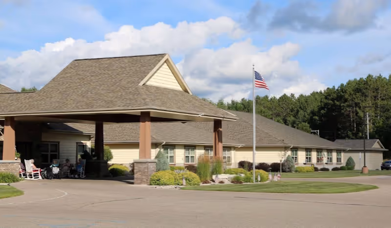 Single-story beige senior living building with a covered porte-cochère entrance, an American flag on a pole, and landscaped grounds.