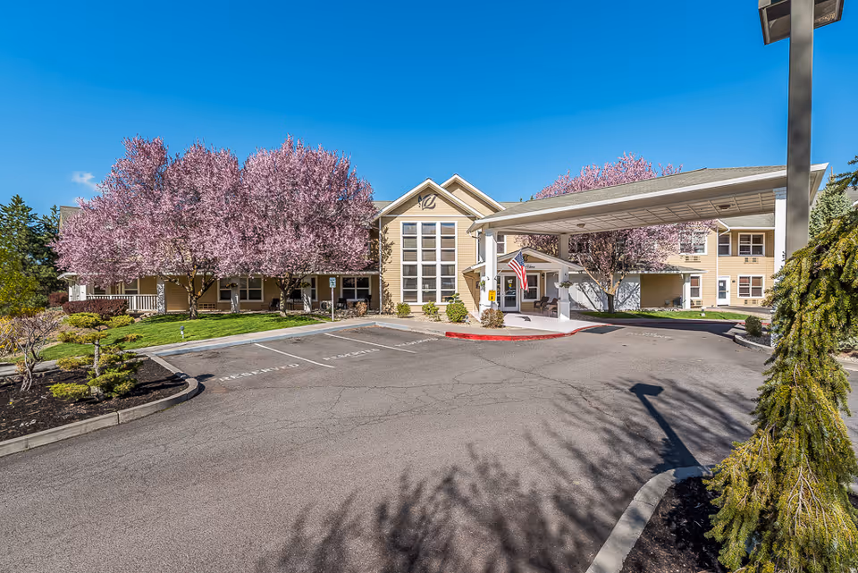 Front exterior of a senior living building with a covered driveway, blooming pink trees, and an American flag.