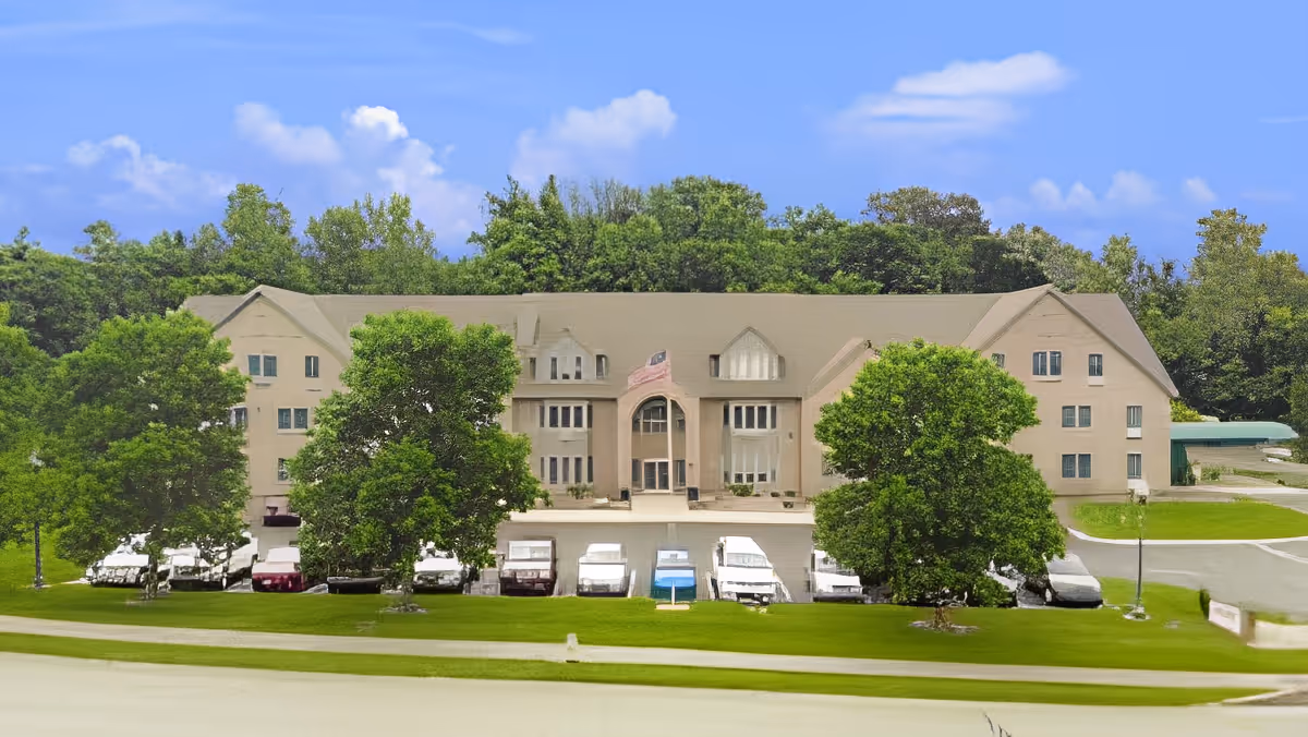 Front exterior of a multi-story beige senior living building with trees, parked cars, and an American flag